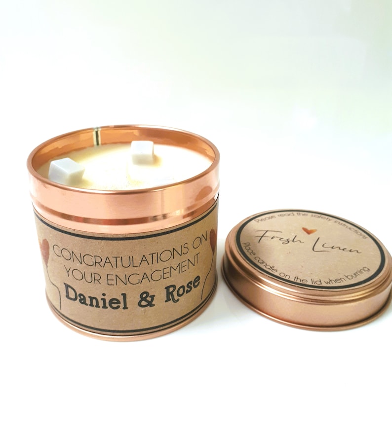May include: A copper candle tin with a brown label that reads "Congratulations on your engagement Daniel & Rose". The candle is white and has three white wax cubes on top. The lid of the tin is copper and has a brown label that reads "Fresh Linen" with a heart symbol. The label also says "Place candle on the lid when burning."