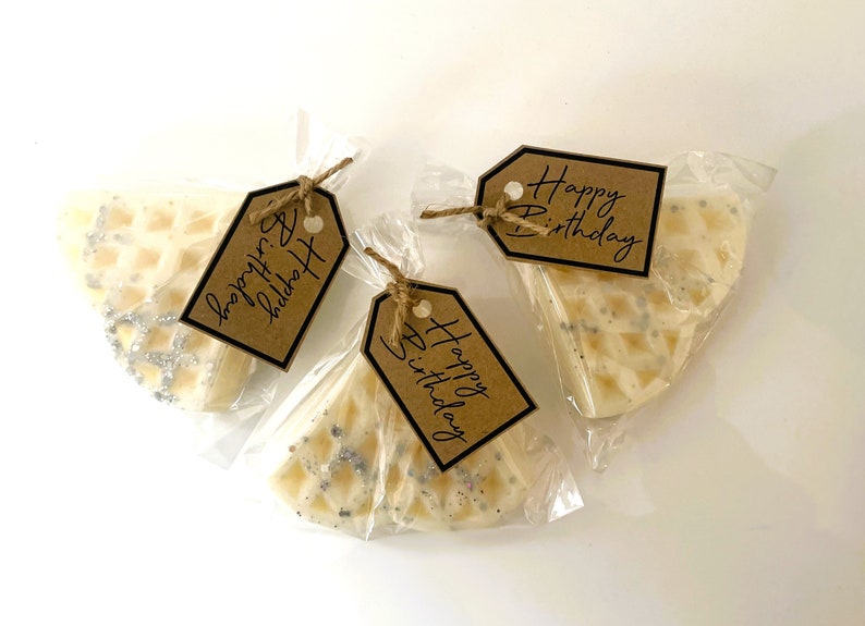 Peut inclure: Trois petites bougies blanches en forme de gaufre avec un motif de paillettes argent&eacute;es, chacune emball&eacute;e dans un sac en plastique transparent avec une &eacute;tiquette en papier brun. Les &eacute;tiquettes portent le texte "Happy Birthday" &eacute;crit en noir.