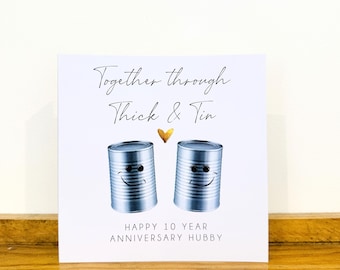 May include: A white greeting card featuring two silver tin cans with cartoon faces, a small gold heart, and the text Together through Thick & Tin and HAPPY 10 YEAR ANNIVERSARY HUBBY printed in black ink on the front of the card.