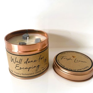 May include: A copper candle tin with a brown label that reads "Well done for Escaping Happy Retirement Steve". The candle is white with gray decorative pieces and has a wick. The lid of the tin is copper and has a brown label that reads "Please read the safety instructions Fresh Linen Place candle on the lid when burning."