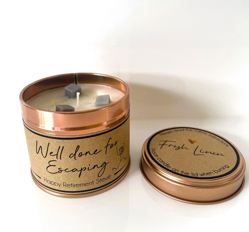 May include: A copper candle tin with a brown label that reads "Well done for Escaping Happy Retirement Steve". The candle is white with gray decorative pieces and has a wick. The lid of the tin is copper and has a brown label that reads "Please read the safety instructions Fresh Linen Place candle on the lid when burning."