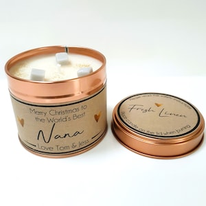 May include: A copper candle tin with a brown label that reads "Merry Christmas to the World's Best Nana - Love Tom & Jess". The candle is white and has three white wax cubes on top. The lid of the tin is copper and has a brown label that reads "Fresh Linen - Place candle on the lid when burning".