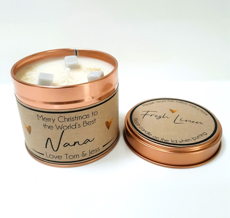 May include: A copper candle tin with a brown label that reads "Merry Christmas to the World's Best Nana - Love Tom & Jess". The candle is white and has three white wax cubes on top. The lid of the tin is copper and has a brown label that reads "Fresh Linen - Place candle on the lid when burning".