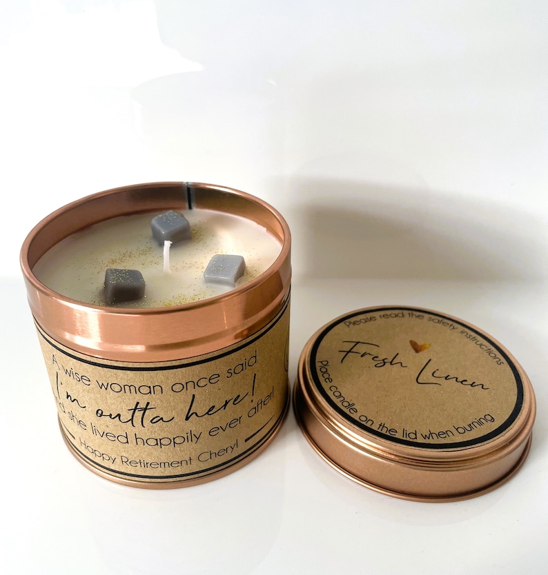 May include: A copper candle tin with a white candle and gray decorative pieces. The label reads "A wise woman once said I'm outta here! She lived happily ever after. Happy Retirement Cheryl." The lid is off and shows a brown label with the text "Please read the safety instructions Fresh Linen Place candle on the lid when burning."