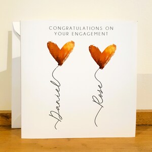 May include: Congratulations on your engagement card with two orange watercolor hearts and the names Daniel and Rose written underneath.