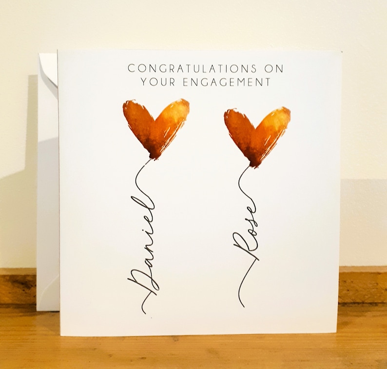 May include: Congratulations on your engagement card with two orange watercolor hearts and the names Daniel and Rose written underneath.