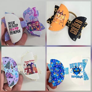 May include: A collection of handmade hair bows with various designs. One bow features the text "EAT SLEEP K-POP REPEAT". Other bows have cartoon graphics, text, and glitter accents. Colors include white, purple, blue, orange, and black.