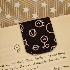 May include: Black fabric bookmark with white geometric patterns. The bookmark is folded and placed on a book with text.