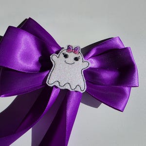 Könnte beinhalten: Eine leuchtend violette Haarschleife aus Satin mit einer zentralen, weiß glitzernden Geisterapplikation. Der Geist hat gestickte schwarze Augen, einen lächelnden Mund und eine kleine rosa Schleife auf dem Kopf. Die Schleife hat zwei lange Bänder.