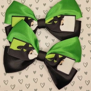 May include: Two green and black hair bows featuring a cartoon image of a black cat with green eyes. The cat is wearing a white shirt and has a green background.