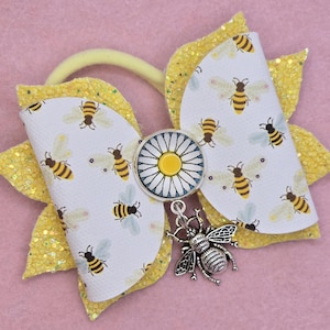 May include: A yellow and white hair bow with a bee theme. The bow features a white background with bee illustrations, a daisy centre, and a bee charm. The bow is attached to a yellow elastic band and has yellow glitter accents.