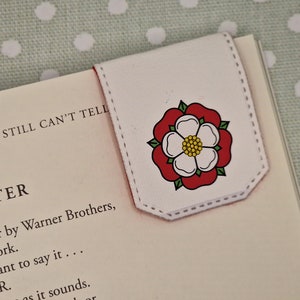 May include: White bookmark with a red and white rose design. The bookmark has a stitched edge and is folded to create a tab.
