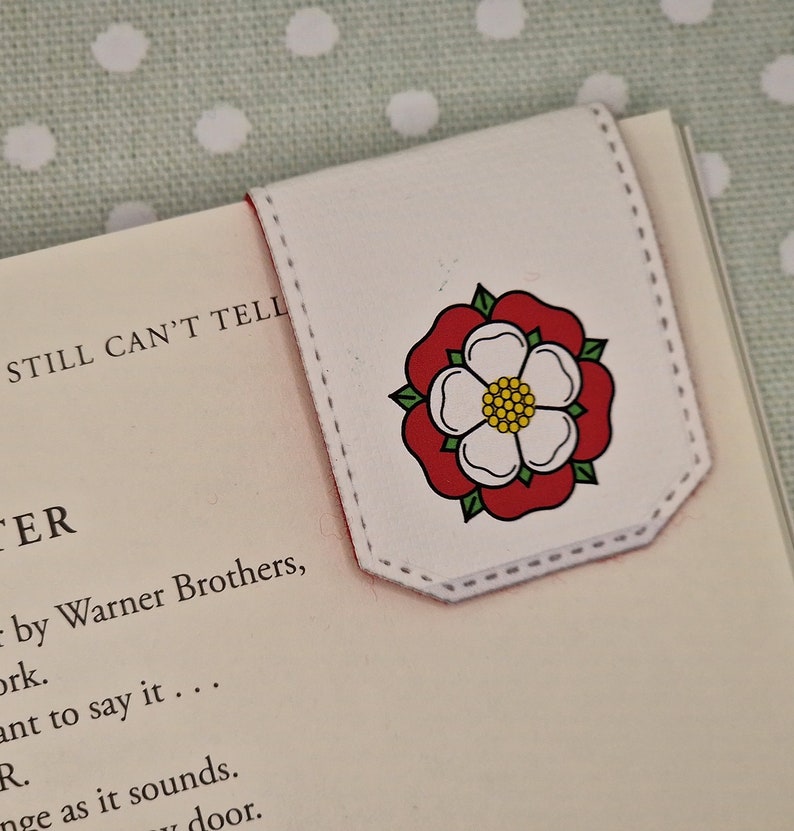 May include: White bookmark with a red and white rose design. The bookmark has a stitched edge and is folded to create a tab.