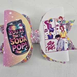 May include: A hair bow featuring a glittery, multi-colored base. The left side displays a soda can graphic with the text "YOU'RE MY SODA POP!" and the right side shows an illustration of five characters with the text "SUIT BOYS" in a grid.