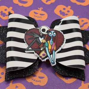 May include: A black and white striped bow with black glitter accents features a heart-shaped image of Jack Skellington and Sally from The Nightmare Before Christmas.