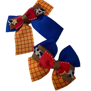 May include: Two hair bows made of grosgrain ribbon featuring a multi-coloured pattern of yellow with red grid lines, solid royal blue, brown, and white with black cow spots. Each bow is accented with a small, gold glittery star-shaped charm.