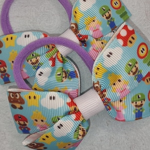 Puede incluir: Dos gomas para el pelo de color morado con un lazo azul y blanco. El lazo está hecho de cinta con un patrón repetido de personajes de Mario, incluyendo Mario, Luigi, Peach, Toad y Goomba.