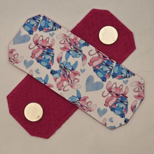 Puede incluir: Una correa de tela blanca con un patrón de dibujos animados rosa y azul de dos personajes que se toman de la mano. La correa tiene dos piezas de fieltro rosa con broches de metal dorado en cada extremo.