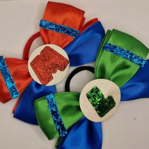 Peut inclure: Accessoires pour cheveux avec des nœuds en satin rouge, bleu et vert, ornés de paillettes. Chaque nœud comporte un centre circulaire avec un motif scintillant. Fixés à des élastiques noirs, adaptés à diverses coiffures.