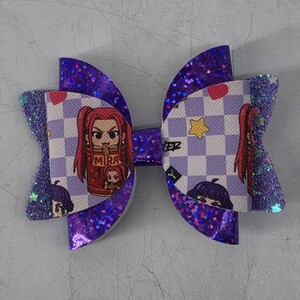 May include: A hair bow featuring layers of purple glitter fabric and a white and light purple checkered pattern. The pattern includes cartoon characters with red hair, a red cup labeled MIRA, red hearts, and yellow stars. The bow has a center knot.
