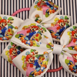 Puede incluir: Un lazo blanco con una banda elástica rosa. El lazo está decorado con una impresión colorida de los personajes de Mario y Luigi del videojuego Super Mario Bros.