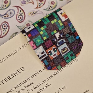 May include: A colourful bookmark with a pixelated design on a black background. The bookmark is made of leather and has a textured surface.