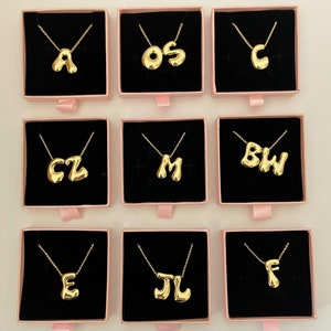 May include: Ten small boxes with black velvet interiors, each containing a gold-plated necklace with a different letter of the alphabet. The letters are in a bubbly, irregular font.
