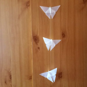 Könnte beinhalten: Drei weiße Papier-Origami-Schmetterlinge, die an einer Schnur an einer Holzwand hängen.