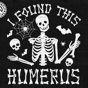 May include: A white graphic design on a black background featuring a skeleton holding a bone, surrounded by a spider web, spiders, bats, and two skulls. The text reads I FOUND THIS HUMERUS, with the letters styled to resemble individual bones.