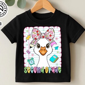 Pode incluir: Camiseta preta com um desenho de ganso de desenho animado, usando um la&ccedil;o e rodeada por imagens escolares. As palavras "second grade" s&atilde;o exibidas em letras coloridas. A camiseta est&aacute; pendurada em um cabide de madeira.