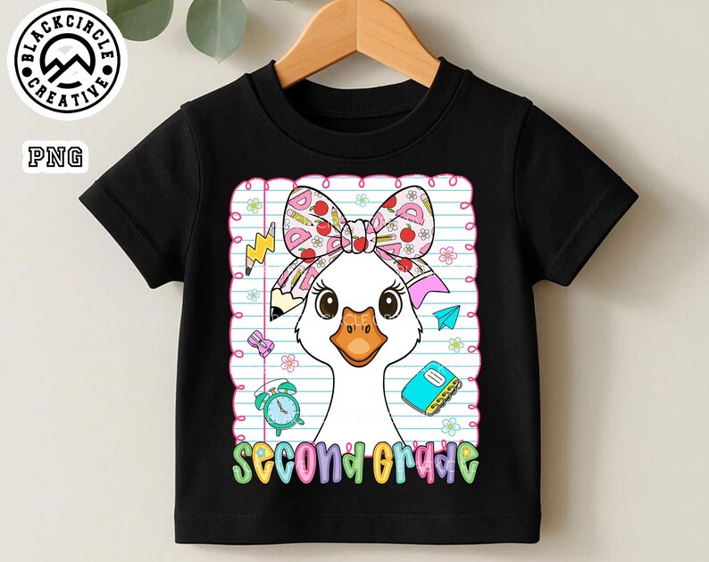Pode incluir: Camiseta preta com um desenho de ganso de desenho animado, usando um la&ccedil;o e rodeada por imagens escolares. As palavras "second grade" s&atilde;o exibidas em letras coloridas. A camiseta est&aacute; pendurada em um cabide de madeira.