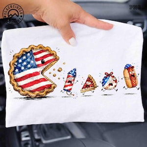 May include: A white t-shirt featuring a graphic of a large pie with an American flag pattern chasing a firework, a slice of pie, a decorated ball, and a hot dog. The characters have wide eyes and open mouths, appearing to run away in fear.