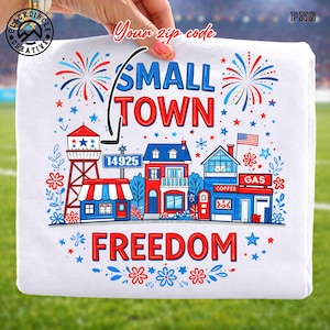 May include: White t-shirt featuring a colorful illustration of a small town with a water tower, houses, and businesses. The text "SMALL TOWN FREEDOM" is displayed in red and blue, with the zip code "14925" included.