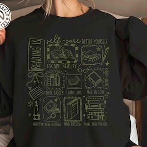 May include: Black sweatshirt with a green graphic design featuring book-related illustrations and text. The text includes phrases like "Reading Is", "Self-Care", "Escape Reality", and "Discover New Worlds".