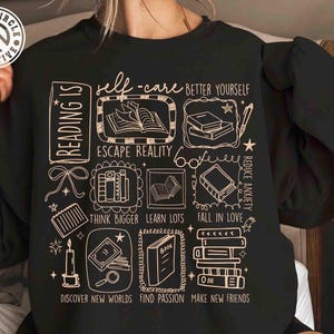 May include: Black sweatshirt with a cream-colored graphic featuring book illustrations and phrases like "self-care," "escape reality," and "discover new worlds." The design includes a logo in the upper left corner.