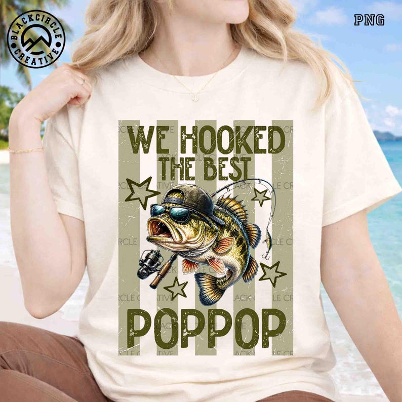 Pode incluir: Camiseta creme com um gr&aacute;fico de um peixe usando &oacute;culos de sol e bon&eacute;, com o texto "WE HOOKED THE BEST POPPOP". O design inclui estrelas e listras verticais em tons de verde e marrom. A camisa tem mangas curtas.
