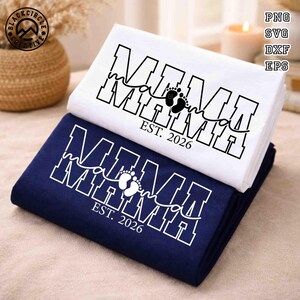 Op de afbeelding: Twee gevouwen stoffen items, één wit en één marineblauw, op elkaar gestapeld. Elk item heeft het woord "MAMA" in vette blokletters, met een script "ma" in het midden, een voetafdruk en de tekst "EST. 2026".