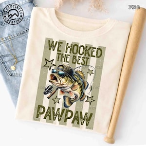 Pode incluir: Camiseta creme com um gr&aacute;fico de um peixe usando &oacute;culos de sol e bon&eacute;, segurando uma vara de pescar. O texto diz "WE HOOKED THE BEST PAWPAW" em verde oliva. Um bast&atilde;o de beisebol de madeira est&aacute; ao lado.