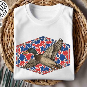 May include: White t-shirt featuring a graphic of a flying duck with outstretched wings. The duck is set against a red, white, and blue camouflage pattern within a hexagonal frame. The shirt is folded and placed in a woven basket.
