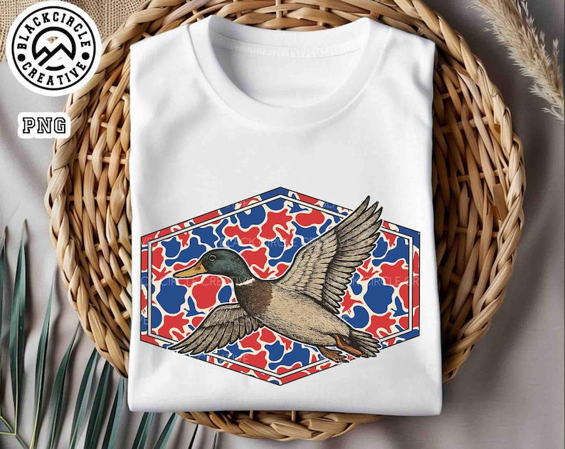 May include: White t-shirt featuring a graphic of a flying duck with outstretched wings. The duck is set against a red, white, and blue camouflage pattern within a hexagonal frame. The shirt is folded and placed in a woven basket.