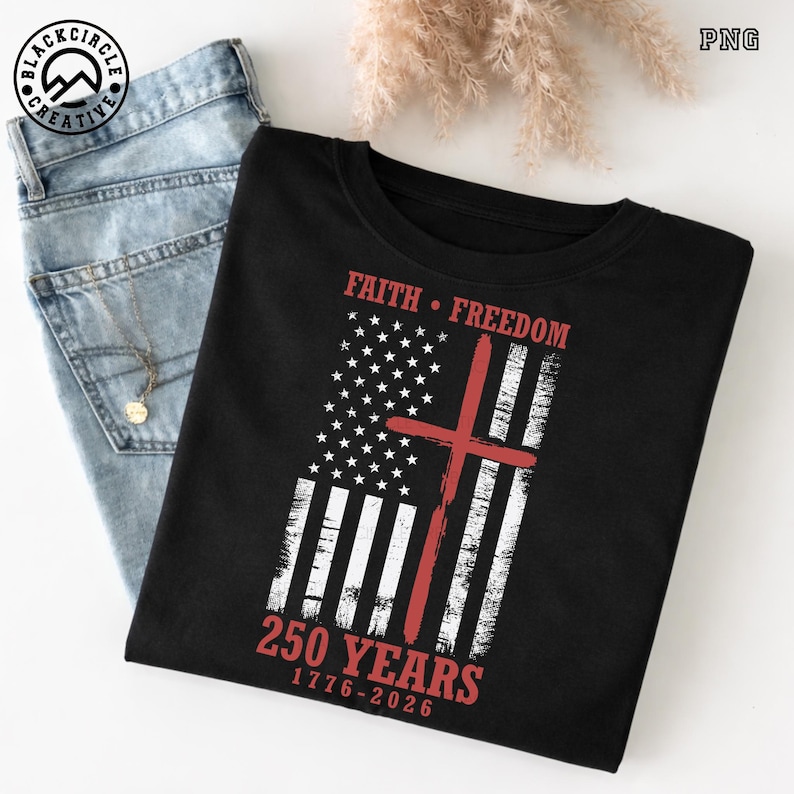 Puede incluir: Camiseta negra con un dise&ntilde;o desgastado de la bandera estadounidense y una cruz roja. Las palabras "Faith" y "Freedom" est&aacute;n encima de la bandera, y "250 YEARS 1776-2026" debajo. La camiseta se muestra con vaqueros azul claro.