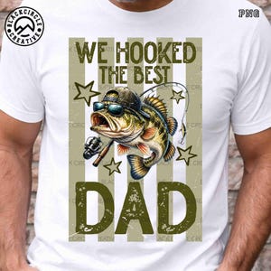 Pode incluir: Camiseta branca com um gr&aacute;fico de um peixe usando &oacute;culos de sol e bon&eacute;, segurando uma vara de pesca. O texto "WE HOOKED THE BEST DAD" &eacute; verde-oliva. O design inclui estrelas e listras verticais.