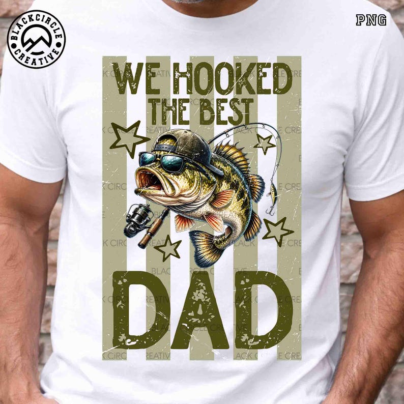 Pode incluir: Camiseta branca com um gr&aacute;fico de um peixe usando &oacute;culos de sol e bon&eacute;, segurando uma vara de pesca. O texto "WE HOOKED THE BEST DAD" &eacute; verde-oliva. O design inclui estrelas e listras verticais.