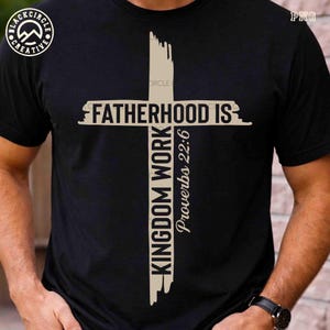 May include: Black t-shirt featuring a beige cross design with the text "FATHERHOOD IS" across the top and "KINGDOM WORK Proverbs 22:6" vertically and on the right side. The shirt has a crew neck and short sleeves.