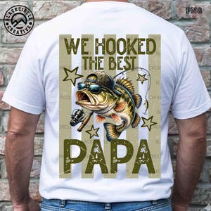 Pode incluir: Camiseta branca com um gr&aacute;fico de um peixe usando &oacute;culos de sol e bon&eacute;, segurando uma vara de pescar. O texto "WE HOOKED THE BEST PAPA" &eacute; impresso em verde oliva. A camiseta tem um visual desgastado.