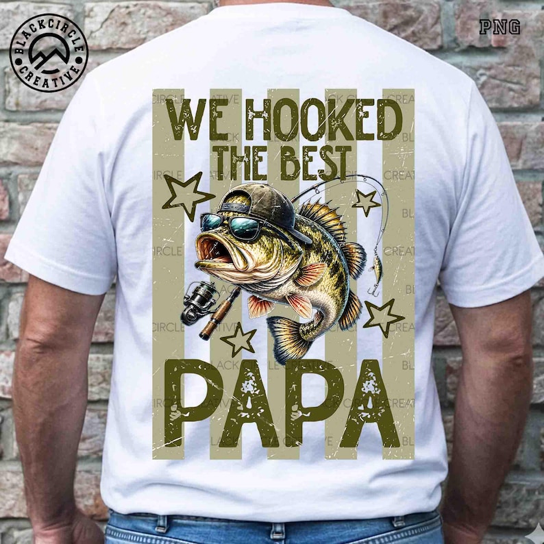 Pode incluir: Camiseta branca com um gr&aacute;fico de um peixe usando &oacute;culos de sol e bon&eacute;, segurando uma vara de pescar. O texto "WE HOOKED THE BEST PAPA" &eacute; impresso em verde oliva. A camiseta tem um visual desgastado.