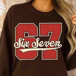 May include: A brown sweatshirt featuring the number "67" in a bold, red, collegiate-style font with a white outline. Below the number, the words "Six Seven" are written in a cursive font with a white banner. The design is centered on the chest.