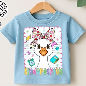 Pode incluir: Camiseta azul claro com um desenho de ganso de desenho animado usando um la&ccedil;o com material escolar. O ganso est&aacute; em frente a um fundo de papel pautado. A palavra "Kindergarten" est&aacute; escrita em letras coloridas na parte inferior.