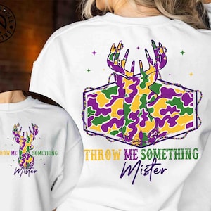 May include: White sweatshirt with a deer head design in purple, green, and yellow camouflage. The text "THROW ME SOMETHING Mister" is printed below the graphic in yellow and purple. The design is repeated on a smaller scale.