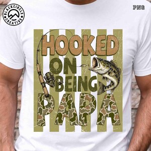 Op de afbeelding: Wit T-shirt met een visserijthema. Het ontwerp toont de tekst "HOOKED ON BEING PAPA" in bruine en camouflage letters, een hengel en een vis met open mond.